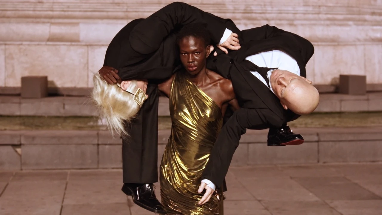 Official Matières Fécales The Ninety Nine Percent film image, showing a runway model carrying two suited figures